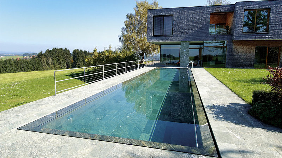 Ein Außenschwimmbecken mit Überlaufrinne. Im Hintergrund: ein Wohnhaus.