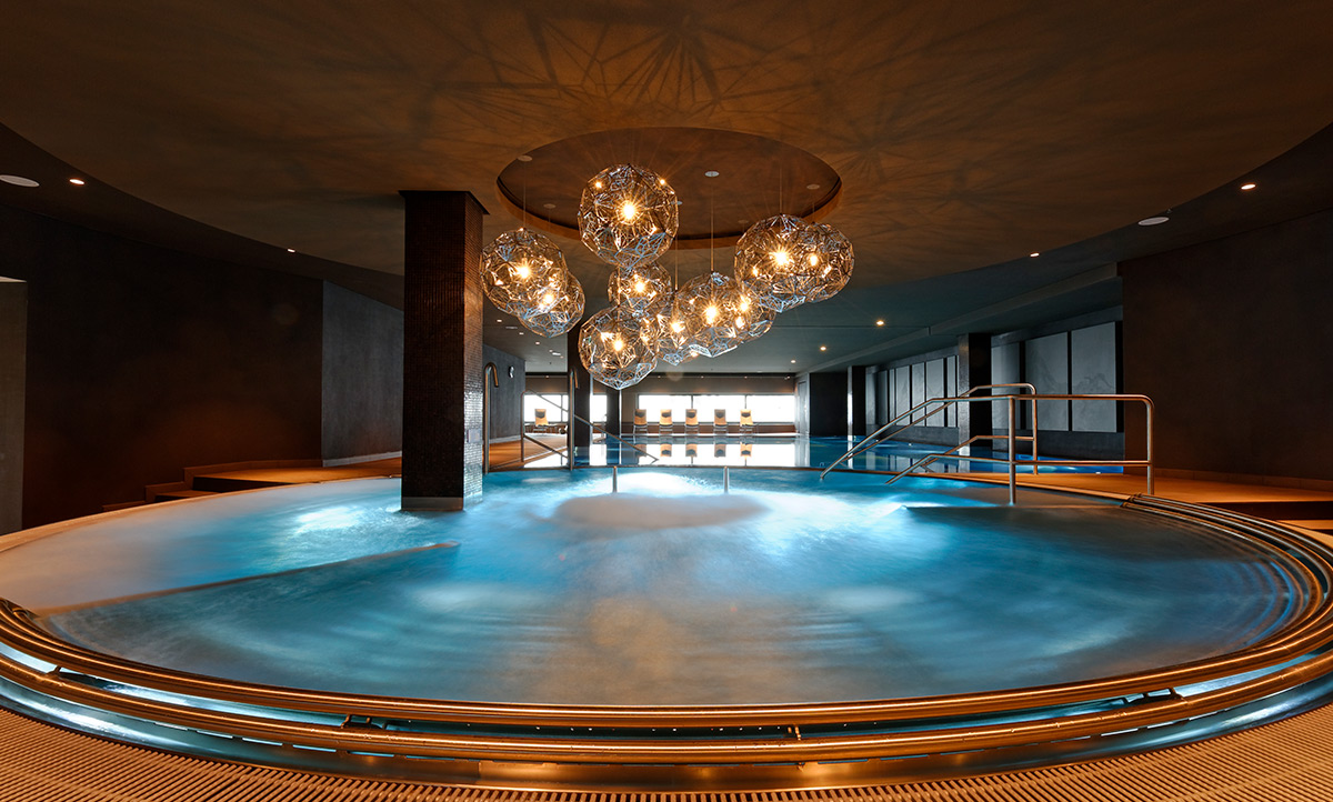 Ein großer Rundpool aus Edelstahl in einem Hallenbad. Über ihm hängen große moderne Leuchten.