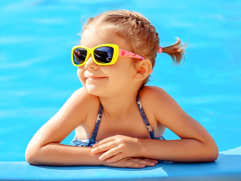 Een meisje met een zonnebril leunt op de rand van het zwembad. 